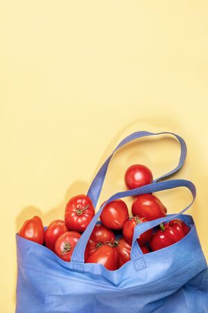 Blue non-woven shopping bag full of fresh red ripe farm tomatos and pepper on yellow background with copy space.の写真素材