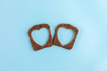 Two rye bread slices with carved holes of heart shape in them on light blue background. Creative minimal concept.の写真素材