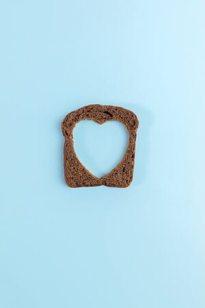 One rye bread slice with carved hole of heart shape in it on light blue background. Creative minimal concept.の写真素材