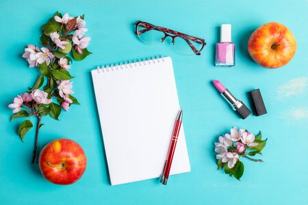Spring turquoise layout with white notepad with pen, women's glasses, pink lipstick and nail polish, two apples and pink Apple tree twigs. Woman home office concept. Top view, flat lay, copy space.の写真素材