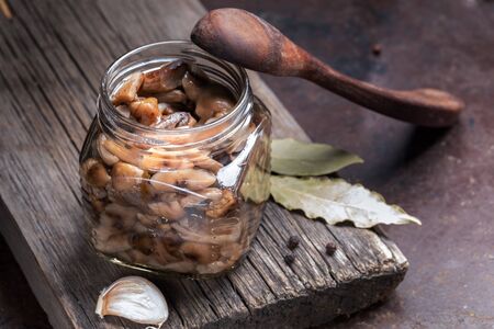 Glass jar with homemade pickled forest mushrooms honey agarics, wooden spoon and spices on old wooden board on rusty metal backdrop. Fermented healthy food. Sustainable food. Close-up.の写真素材
