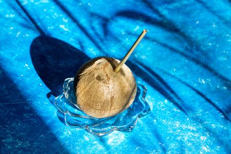 Fresh coconut with bamboo straw inserted in it on glass plate on blue background with shadows and copy space. Zero waste concept. Rejection of plastic drinking straws. Trendy image with hard shadows.の写真素材