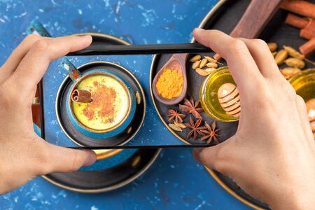 Female hands holding smartphone with photo above of blue cup of traditional indian ayurvedic healthy drink golden turmeric latte milk with ingredients on blue background. Top view. Creative photo.の写真素材
