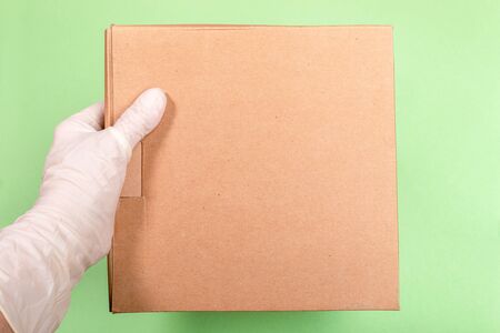 Female hand in white latex glove holding brown cardboard box on light green background. Concept safe...