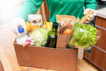 Woman in protective gloves taking food products out of box on wooden table in kitchen. Safe delivery. Contactless home delivery during quarantine. Food donation. Stock for rainy day. Selective focus.の写真素材