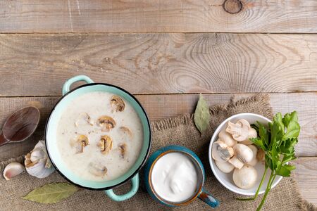 Turquoise ceramic pan with mushroom cream soup, cup with sour cream, bowl with fresh champignons, spoon and spesies on burlap on wooden background. Top view, copy space.の写真素材