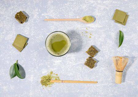 Flat lay arrangement of matcha tea glass, wooden spoons with matcha powder, whisk, leaves,...