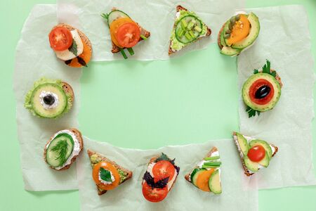 Frame of vegetarian sandwiches with tomatoes, cucumbers, avocado, sun-dried olives, greenness and cheese laid out in circle on parchment paper pieces on green backdrop. Top view, flat lay, copy space.の写真素材