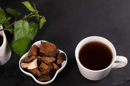 White ceramic cup of healing infusion of birch mushroom chaga, bowl with chaga pieces and birch leaves on black. Chaga mushroom coffee or tea. Trendy healthy superfood for immunity. Copy space.の写真素材