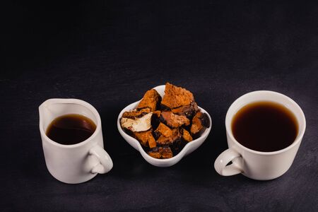 Two white ceramic cups of healing infusion of birch mushroom chaga with chaga pieces in bowl on black backdrop. Chaga mushroom coffee or tea. This drink rich by antioxidants for immunity. Copy space.の写真素材