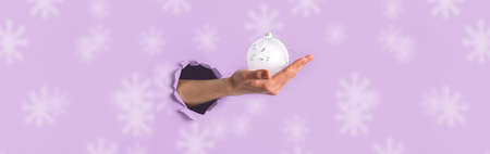 Wide festive Christmas banner with woman hand holding white Christmas ball through torn hole on middle. Pink backdrop with large snowflakes. Creative Xmas invitation. Xmas sale concept. Copy space.の写真素材