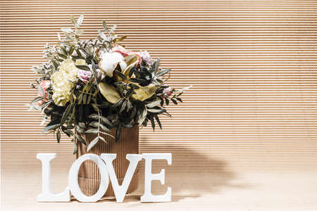 Eco friendly arrangement with flowers bouquet in DIY cardboard vase and white letters LOVE on beige carton backdrop. Modern monochrome greeting card with shadows. Horizontal orientation. Copy space.の写真素材