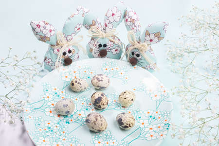 Three DIY textile Easter bunnies standing around plate with spring floral decor with quail eggs on...