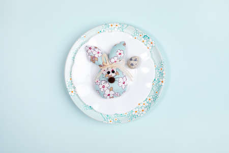 Easter festive minimal layout with one quail egg and one DIY textile Easter bunny on plates on light blue backdrop.の写真素材
