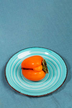 One ugly orange persimmon on turquoise plate on dark turquoise textile napkin. Zero waste food. Fresh healthy organic fruits. Reasonable sustainable consumption. Copy space. Vertical orientation.の写真素材