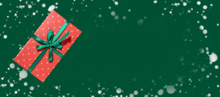 One red rectangular wrapped polka dots gift box decorated green ribbon with bow on deep green backdrop with snow frame. Christmas banner. Xmas festive decorations. Top view. Copy space for text.の写真素材