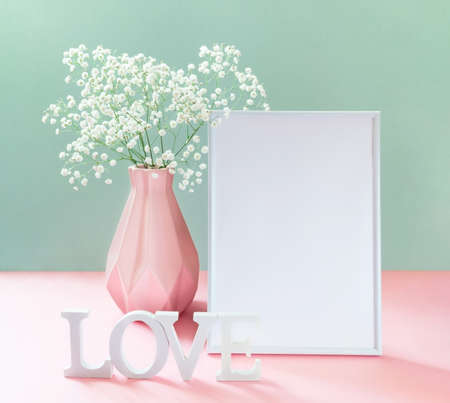 Creative holiday greeting card for Valentine's day, Mother's day, March 8th or Birthday with vase of white gypsophila, empty white frame, white letters LOVE on pastel pink-green. copyspace. Mock up.の写真素材