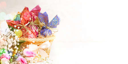 Basket with Easter decorative eggs wrapped in colorful paper with golden ribbon, flowers and color bokeh lights. Festive Easter banner. Holiday decorations. copyspace. horizontal orientation.の写真素材