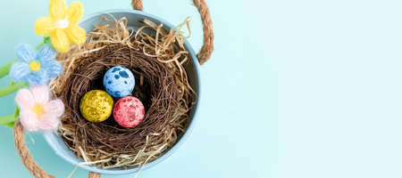 Nest of color painted quail Easter eggs in small blue decorative bucket, knitted flowers on pastel...
