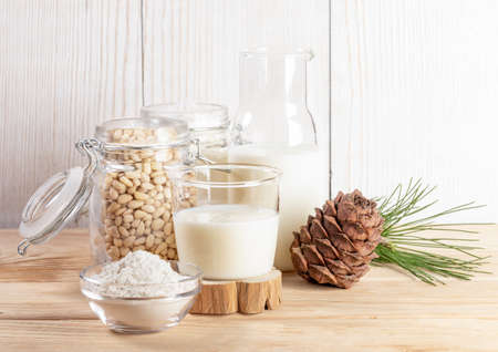 Glass and bottle with vegan cedar milk, flour in bowl and nuts in glass jar, cone, branch on wooden table. Plant based alternative milk. lactose free drink. milk replacer. copyspace. selective focus.の写真素材