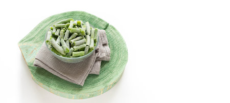 Frozen cut green string beans in glass bowl on gray napkin on green wooden slice on white. organic vegetables. Health food. Sustainable consumption advertising banner. Copy space for text.の写真素材