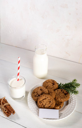 Fresh cookies on round plate, milk in glass and bottle, empty blank note on white wooden table against white wall. Christmas tradition - milk for Santa. Vertical format. Copy space for text.の写真素材