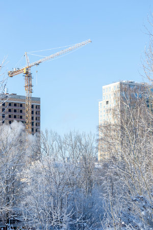 View to construction of multistory new residential house and construction crane in sunny frost winter day. Snowbound trees on foreground. Vertical orientation.の写真素材