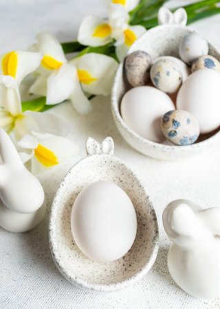 Holiday Easter natural colors eggs in marble bowls with bunny ears, small ceramic bunnies, bouquet of white yellow Iris flowers on white tablecloth. Monochrome Easter image. Close up. Vertical format.の写真素材