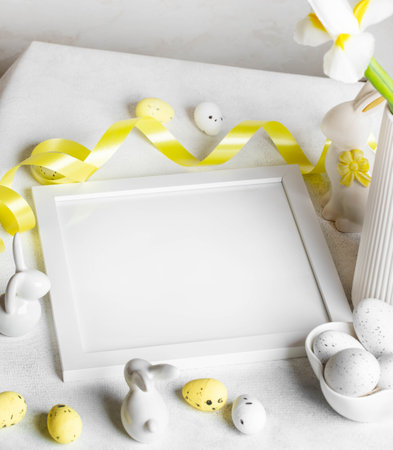 White table with square empty white frame, decorative eggs, flowers, ribbon, ceramic bunnies on white. Easter creative festive card with copy space. Mock up. Template. Holiday image.の写真素材