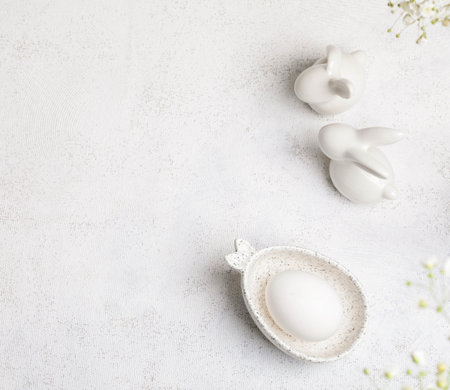 Minimal monochrome white Easter card. Top view One white egg in marble bowl with bunny ears, two ceramic bunnies, Gypsophila flowers on top. Organic Easter. Square format.の写真素材
