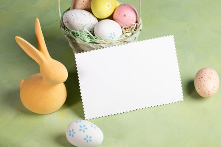 White blank note standing on pastel yellow green backdrop surrounded by decorative color Easter eggs...