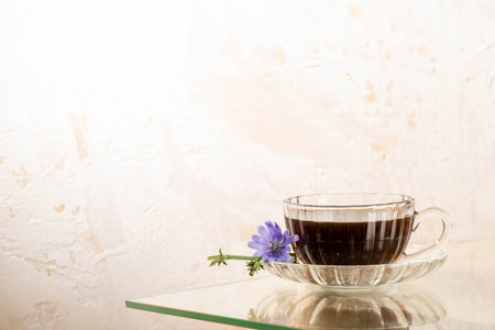 Herbal medicine for human health. Glass cup of chicory tea on saucer with lilac flower on glass table. Light textured wall as background. Chicory tea is prebiotic, good for sleep. Copy space on wall.の写真素材
