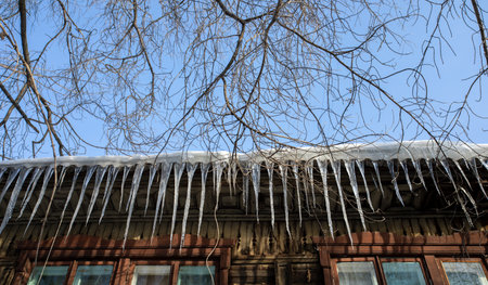 View from bottom to row of spring icicles hanging down from snow covered roof of old wooden house....