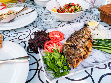 Grilled fish beautifully presented on a marble platter, accompanied by fresh tomatoes, herbs, and greens, set on a patterned tablecloth, creating an inviting dining atmosphereの写真素材