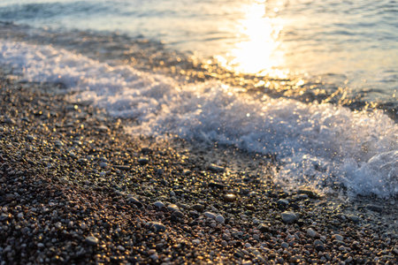 Gentle waves are lapping against a pebbled shore during sunset, with sunlight reflecting on the water, creating a tranquil coastal ambiance filled with natural beauty and peaceの写真素材