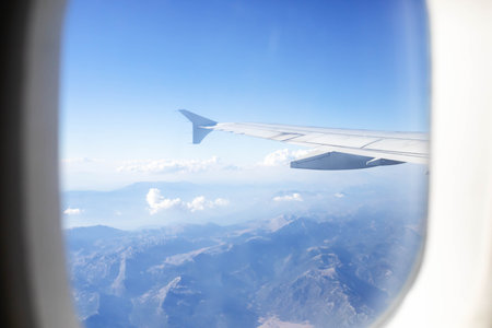 Airplane wing extends into the frame, showcasing breathtaking mountain landscape and fluffy clouds below, illuminated by sunlight, creating a serene atmosphere during the flightの写真素材
