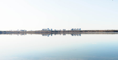 Tranquil scene featuring a still body of water mirroring a distant city skyline under a clear blue sky, creating a peaceful atmosphere with natural beauty and urban elementsの写真素材