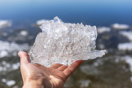 A hand is holding a transparent piece of ice, reflecting sunlight, above a calm water surface. The scene captures the beauty of nature and the intricate details of the ice texture and clarityの写真素材