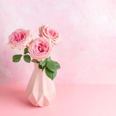 Three pink roses with green leaves are arranged in geometric vase on pink surface, complemented by textured pastel pink background, creating harmonious floral display for romantic holiday cardの写真素材