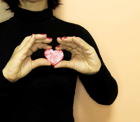 A woman in her 50s wearing black clothes holds a red heart-shaped cookie in her hands on a creamy backgroundの写真素材