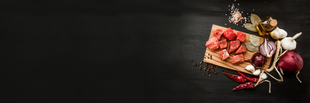 Uncooked fresh diced beef meat with herbs and oil on an old rustic wooden kitchen board over black background. Top view with free space for text. Bannerの写真素材