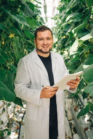 Male agricultural scientist in white lab coat, smiling while holding clipboard, surrounded by lush...