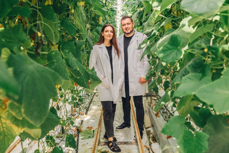 Two individuals in gray lab coats standing among lush green plants in a greenhouse, showcasing...