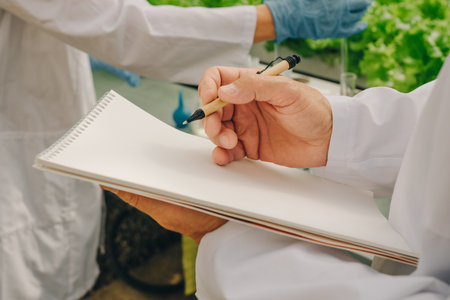 Field study documentation, Researcher meticulously notes plant growth in greenhouse environment, Biologist accurately documenting leaf development with colleague nearby under natural lightingの写真素材