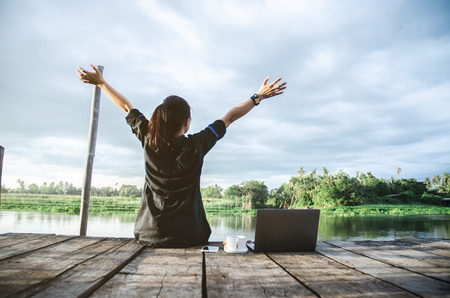 Asian women sitting at the waterfront in the morning.Lady sipping coffee, bread With a notebook computer At the waterfront in the morning.の写真素材
