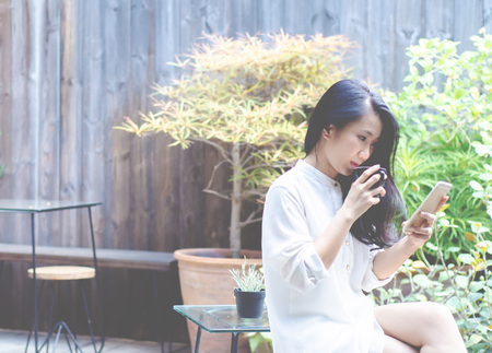 Asian women wear white shirts. She is drinking coffee in the morning garden.Black coffee mug in hand.In the garden there is a girl sitting.Do not focus on objects.の写真素材