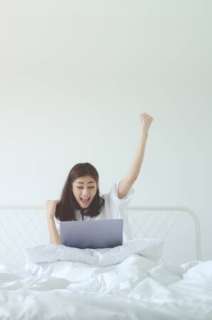 Beautiful Asian woman is smiling.Lady work with laptops on the sofa in the room in the morning.She is happy to get a new job, success, or get good news.In the white room there is a woman inside.の写真素材
