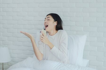 Asian woman wearing glasses Resting by listening to music.White headphones in the hands of girls.In the white bedroom, a lady was resting.Do not focus on the object.の写真素材