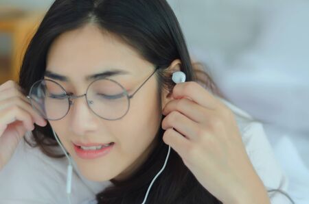 Asian woman wearing glasses Resting by listening to music.White headphones in the hands of girls.In the white bedroom, a lady was resting.Do not focus on the object.の写真素材