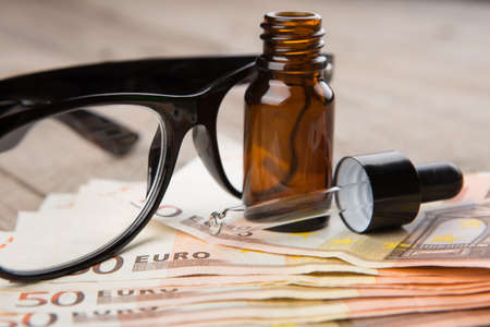 eye care concept - reading glasses, eye drops and money on the wooden deskの写真素材